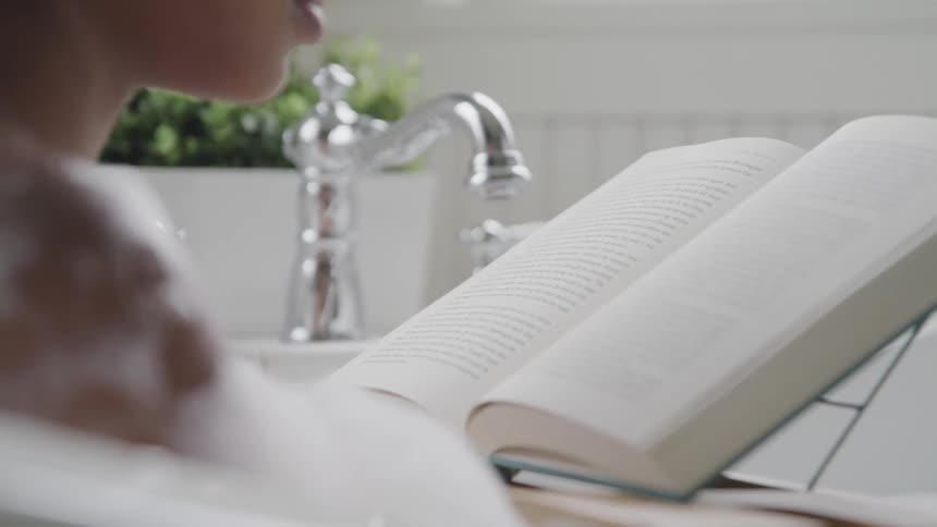 A woman in a bathtub in the bathroom reading a book with plants in the background