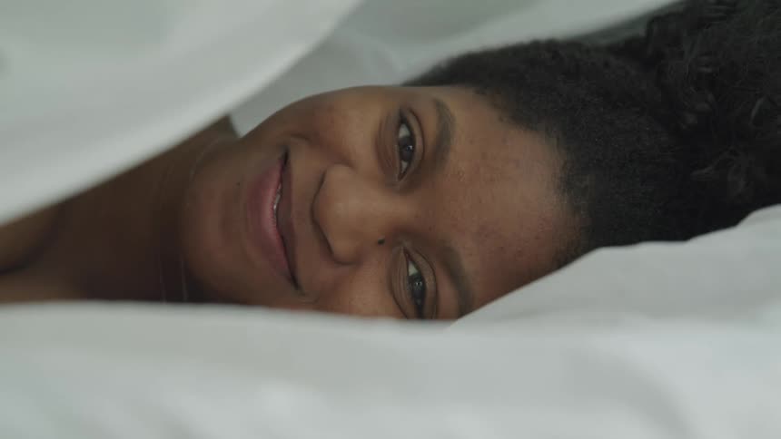 A young woman lying under the covers on a bed lifts the bed sheet to smile into the camera