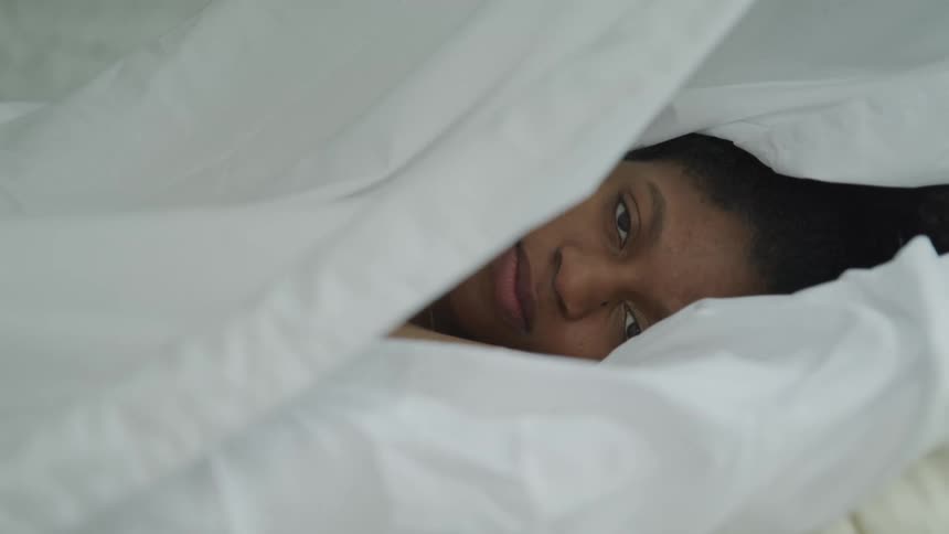 A young woman lying in bed covers her face with a sheet
