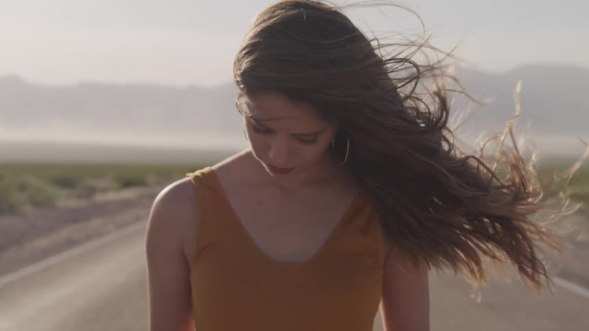 A young woman stands looking downward in the middle of a road running through the countryside as her long hair blows in the wind