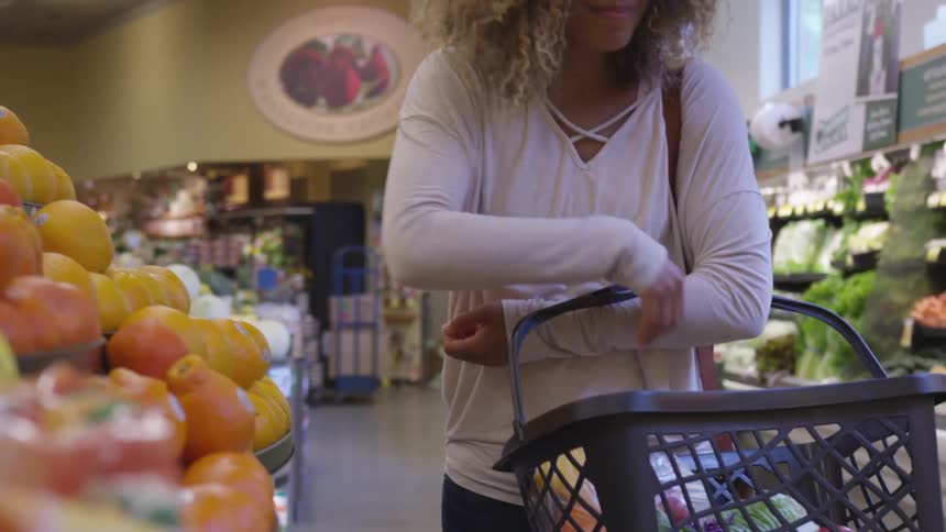 A woman picks up a fruit from a shelf in a grocery store and places it in her shopping basket