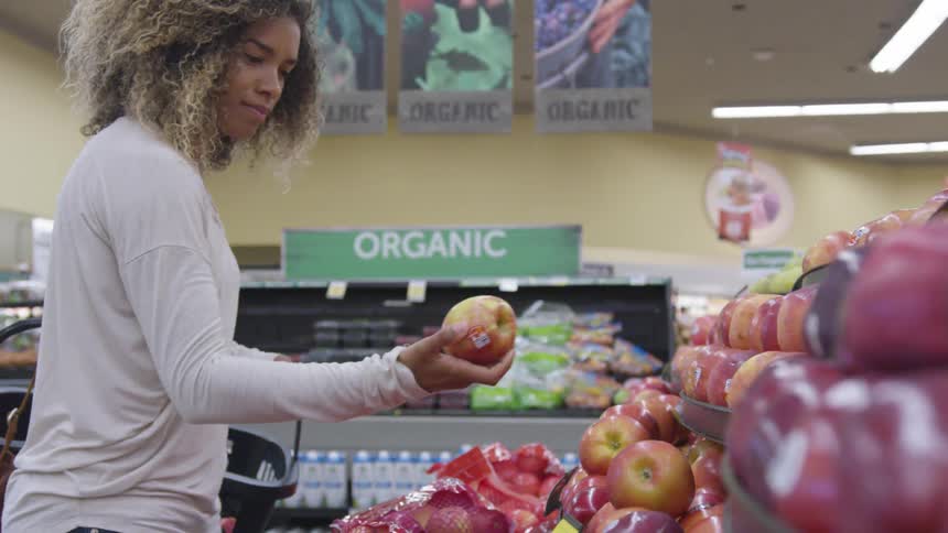 A woman holds a shopping basket and picks up an apple from the store display