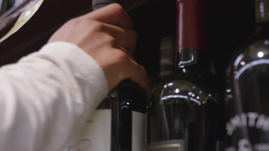 A person reaches up and takes a single bottle of red wine from a shelf in a store