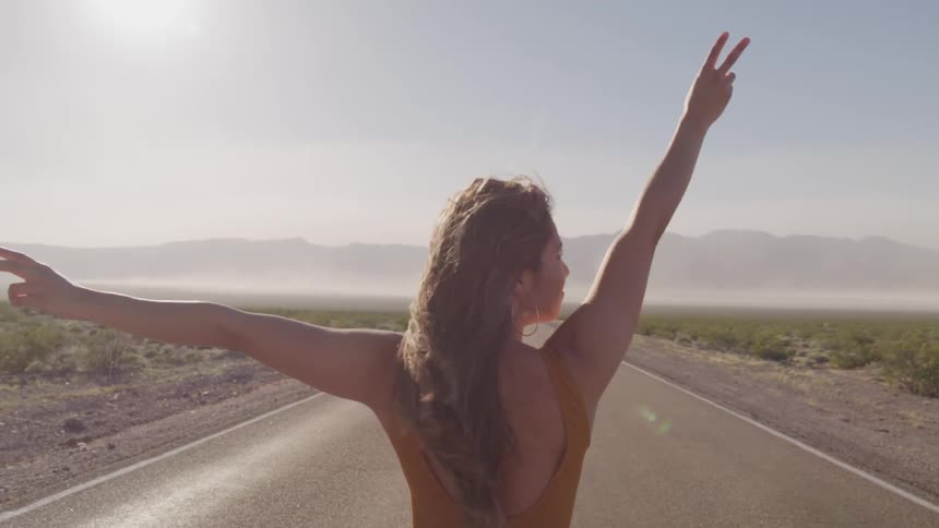 A woman stands on a road through a grassland making the peace sign with her fingers with her arms raised as wind blows through her hairs