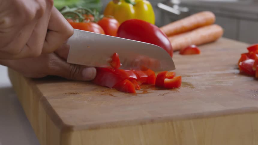 Male using a chefs knife chops slices of a red pepper on a wooden cutting block in the kitchen of their home