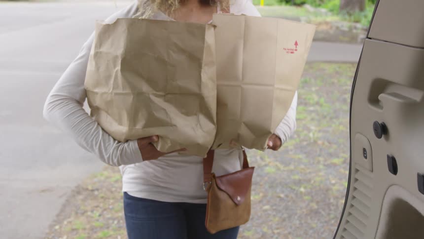 A woman picks up two grocery bags from the back of her vehicle