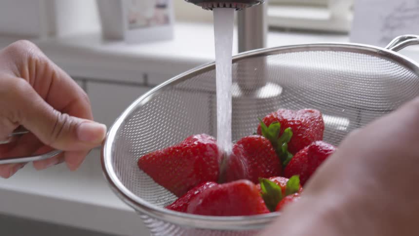 A person washing strawberries in a mesh colander over a kitchen sink
