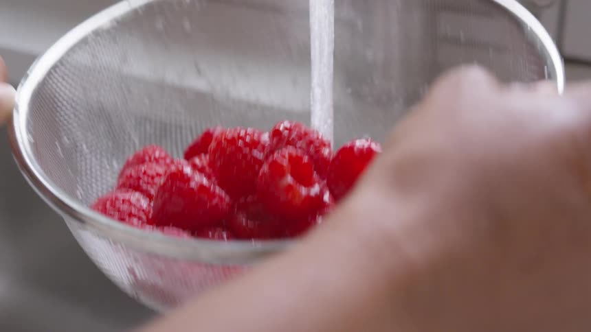 A person rinses raspberries in a metallic colander in the kitchen sink