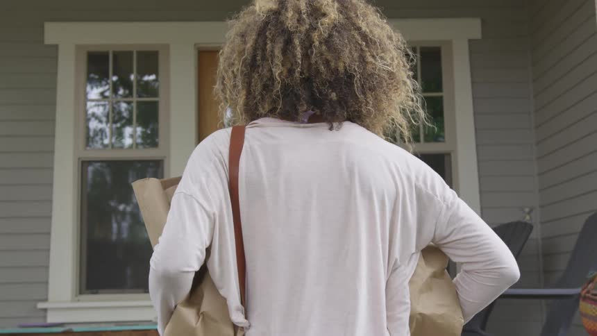 A woman carrying bags of groceries walks up to the front door of a house