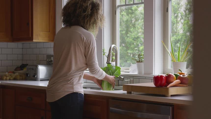 A woman washes a head of lettuce in a kitchen on a sunny day