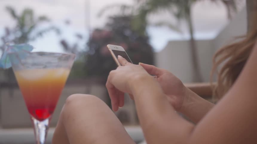 Woman with brown hair and bikini lying by the pool uses a smartphone