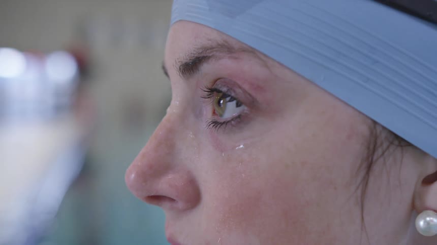 A woman with earrings a swimming cap and goggles raising her head next to a swimming pool