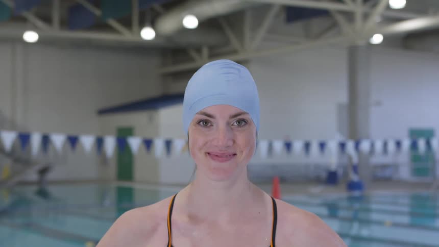 A young female swimmer stands in front of a pool and smiles
