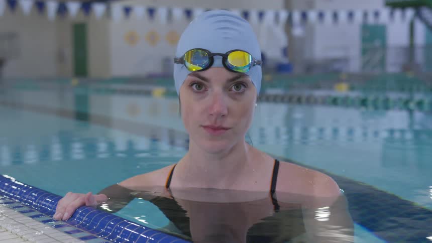 Woman holding edge of indoor swimming pool wearing swim cap and goggles