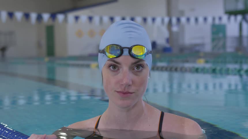 A female swimmer in an indoor swimming pool raises her gaze to look into the camera