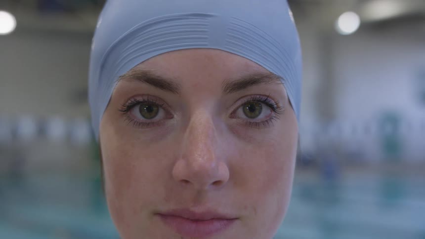 A woman wearing a swimming cap poses in front of an indoor pool