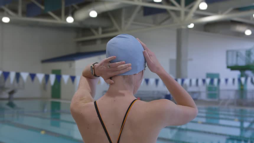 A swimmer adjusts her swim cap as she stands near an empty indoor pool