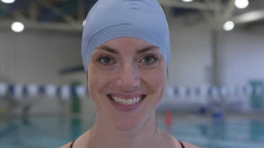 In front of a swimming pool a woman wearing a blue swimming cap raises her head and smiles