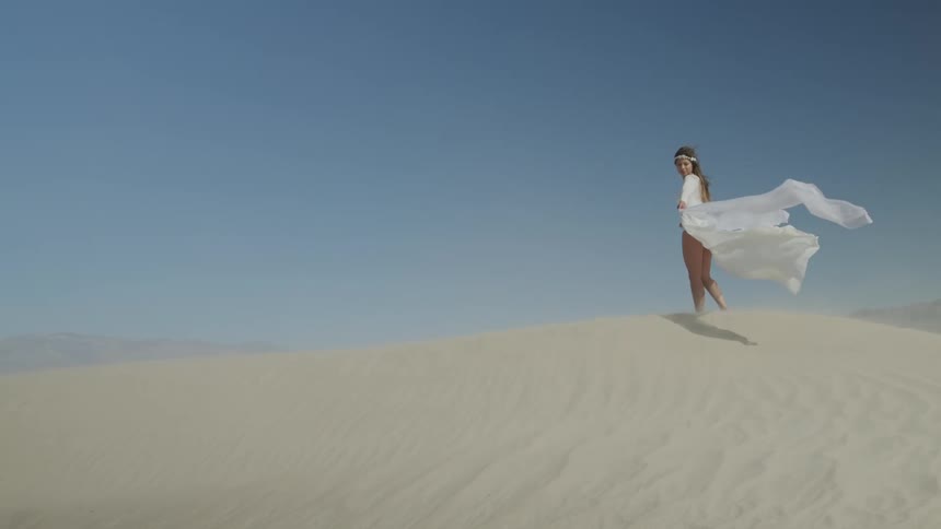 Woman stands on top of sand dune under clear blue sky as wind blows skirt up showing her bare legs