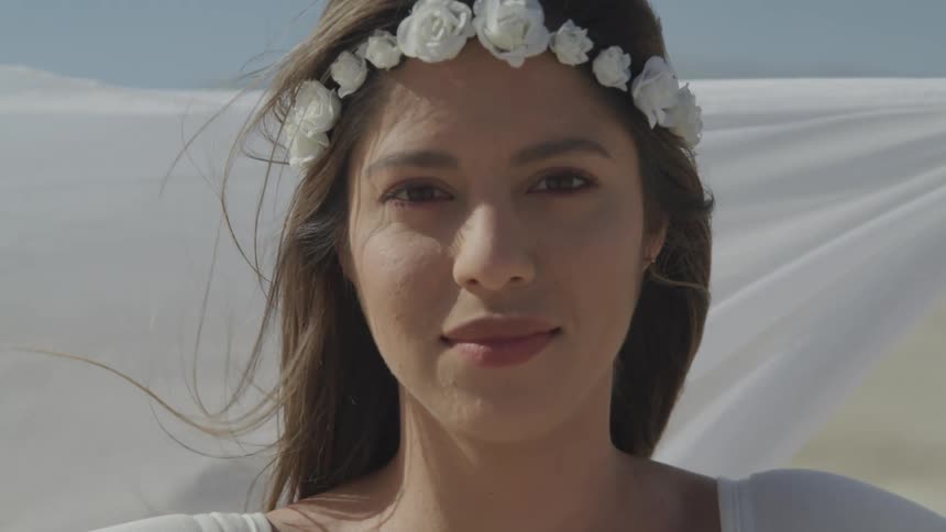 Young woman in a white dress and a wreath of white flowers in her head looks at the camera