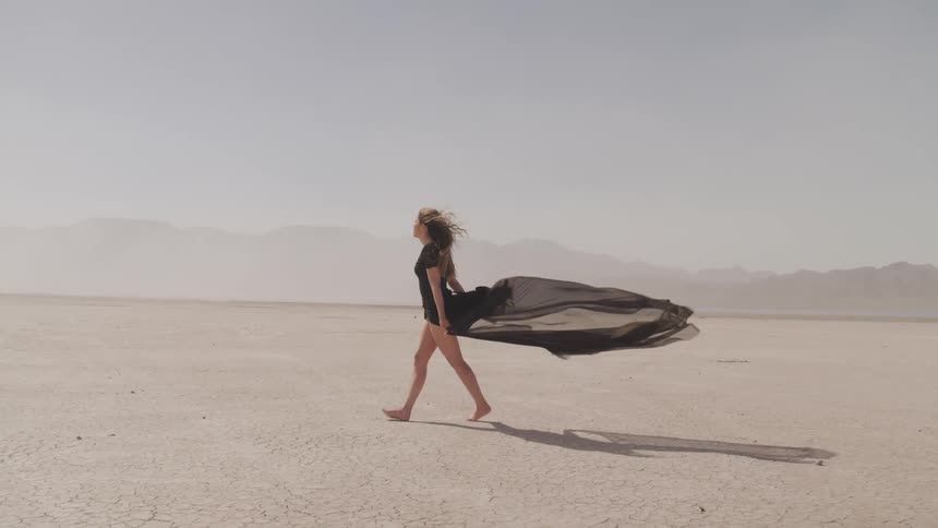 Woman walks across desert as wind blows skirt and filmy cloth behind her