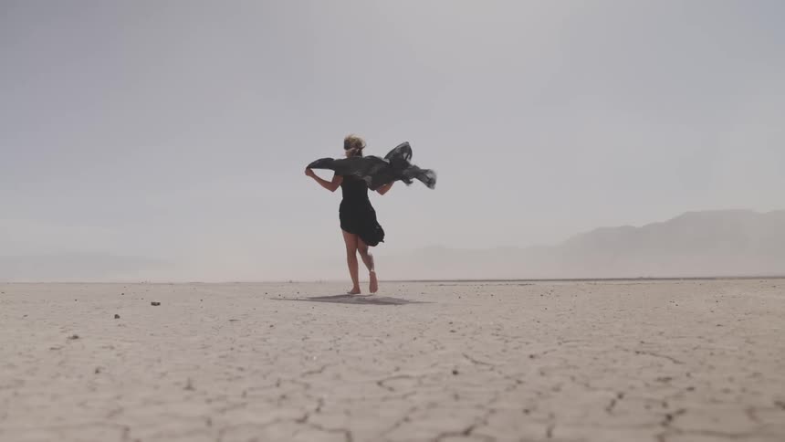 Person walks across arid cracked dessert as dust blown by wind obscures mountains