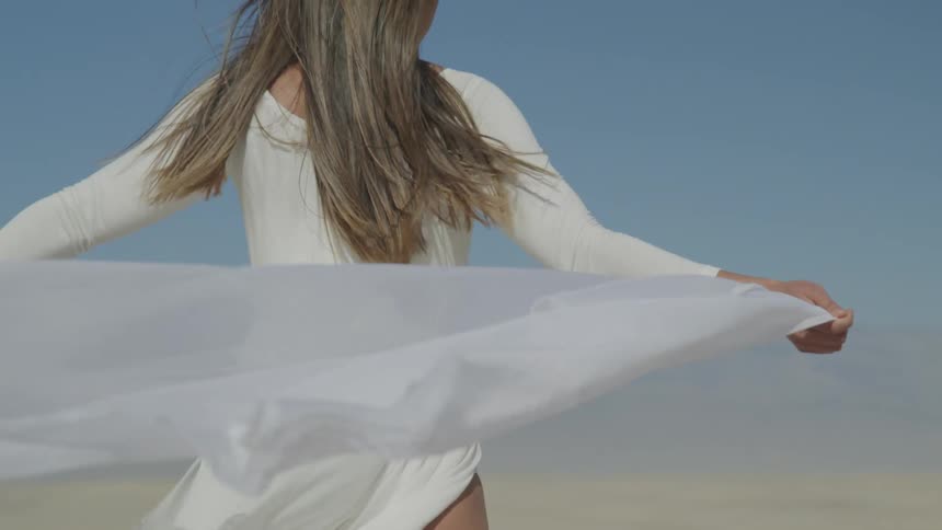 A woman stands on the beach and holds a scarf out behind her as it blows in the wind