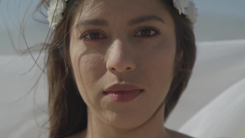 A young woman wearing a wreath of white flowers looks into the camera as wind blows through her hair