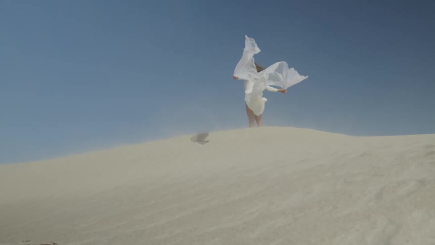 Woman in white gown stands on top of sand dune in breeze