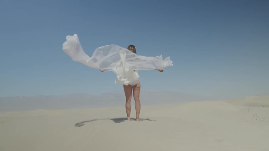 A woman standing on sand with a white dress and white cloth waving in the wind with the sky in the background