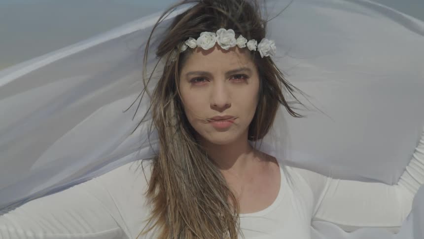 A woman with a white top and a white headband holding white cloth as the wind through it with the sky and clouds in the background