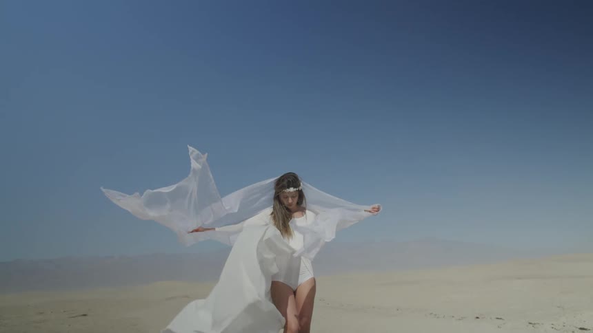A woman stands outdoors in the desert with her arms held wide as her long clothing billows around her in the wind