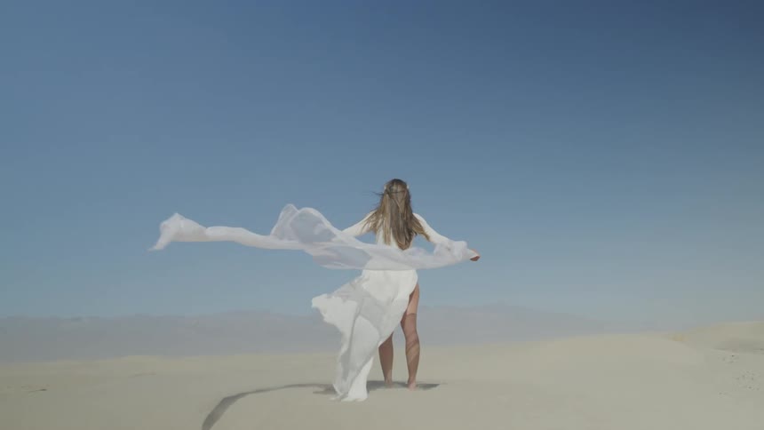 Young woman stands on the desert with the wind blowing her white dress