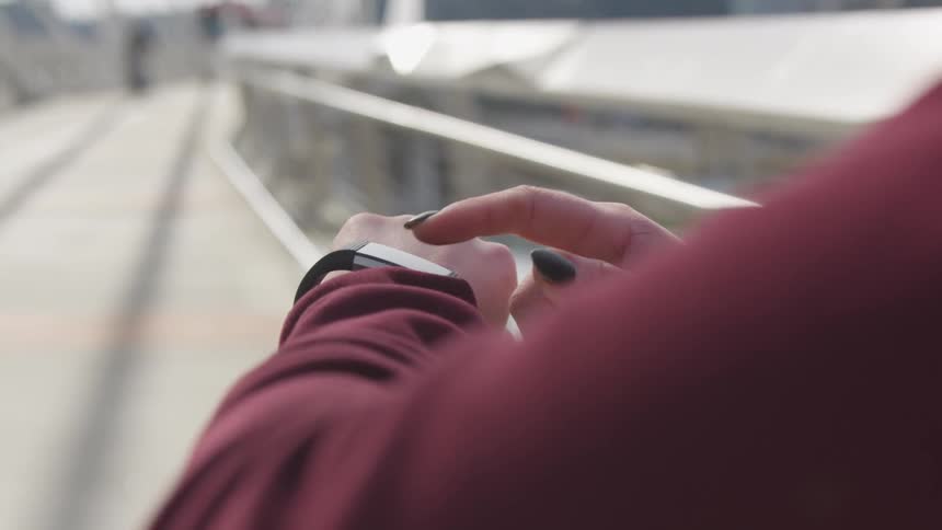 A young wearing black nail polish taps her smartwatch while standing outdoors on a bridge during the day