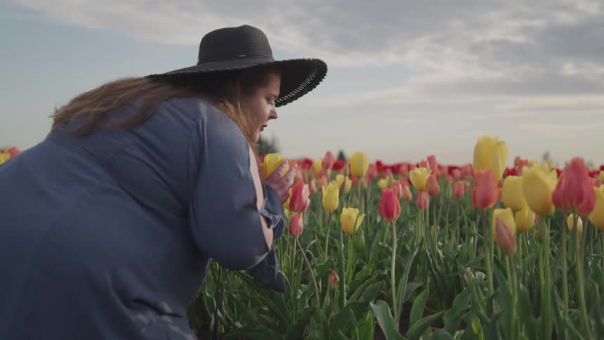 Female standing bend over in a tulip field gently holds a flower in one hand up to her nose while closing her eyes and sniffing in the aroma