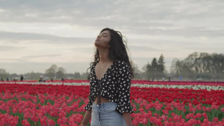 A woman stands in a field of flowers with her eyes closed and head raised towards the sky as others walk in the background