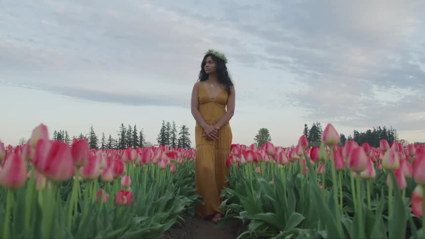 A woman stands in a field of tulips wearing a flower crown and a long yellow dress