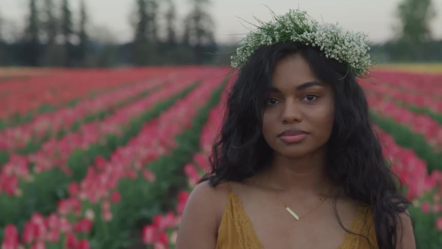 A woman stands in a field of pink tulips with a crown made of small white flowers on her head