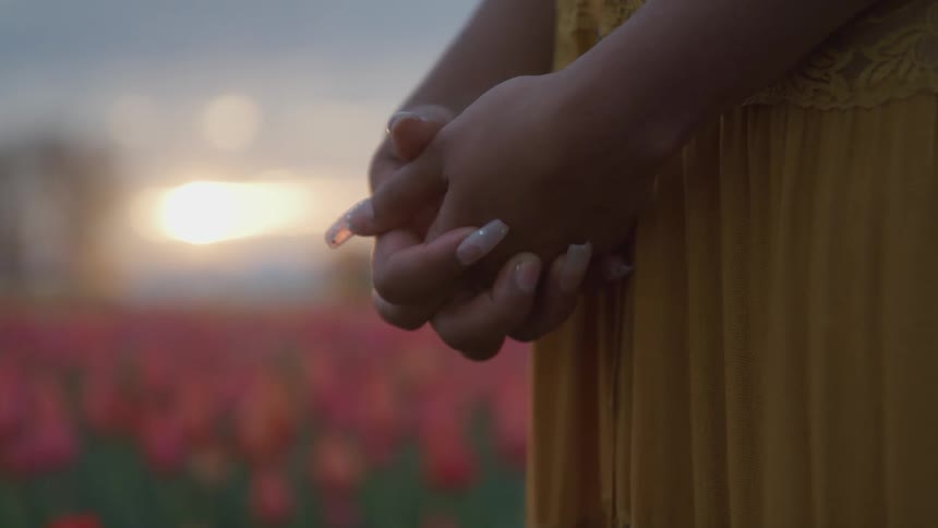 A woman stands in a field of flowers with her hands clasped together in front of her during the day