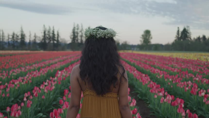 A woman slowly walks in a field of flowers wearing a headdress
