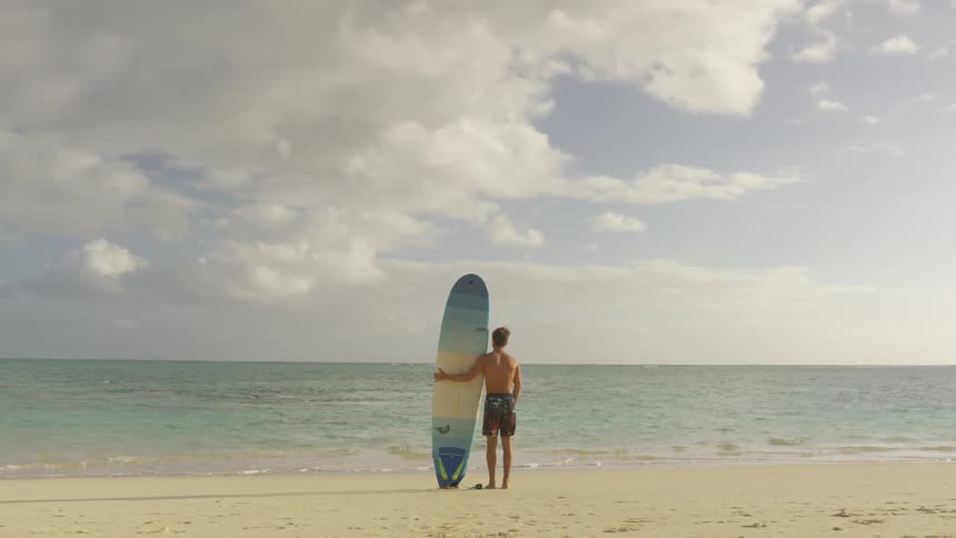 Young man holding surfboard stands on beach looking out to sea