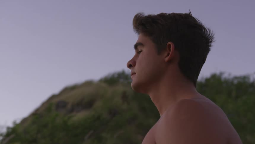 The wind blows through a young caucasian mans hair as he stands shirtless outdoors during the day