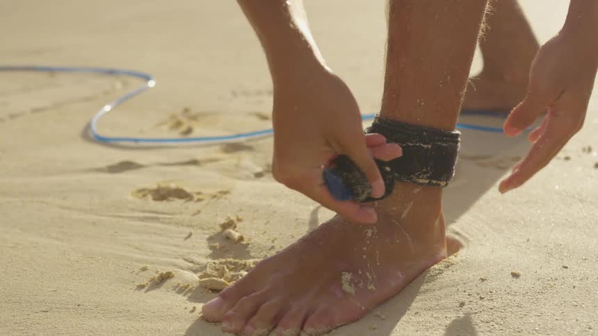 A surfer attaches a surf board leash to their ankle while standing on a beach on a sunny day