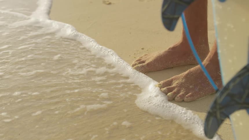 A man stands on the beach with a surfboard as waves roll over his toes