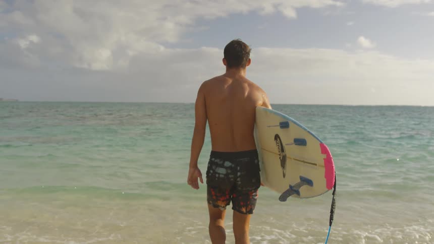 A surfer carries his surfboard under one arm as he walks across a beach towards the ocean