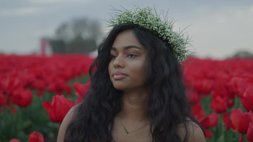 A young woman standing in a field of red flowers with a wreath on her head turns to look into the camera