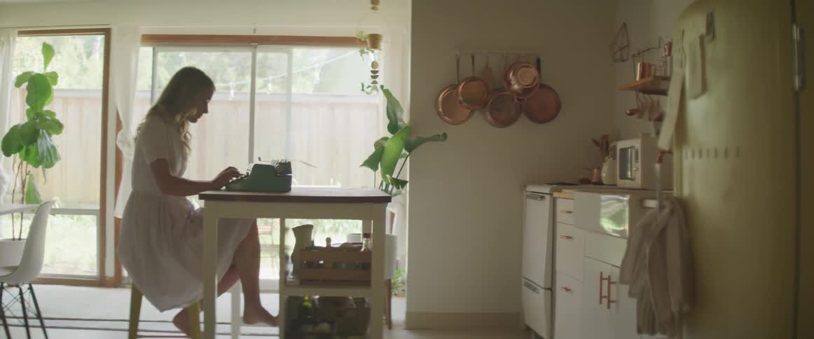 A barefoot woman in a dress sits at a kitchen table typing on a typewriter
