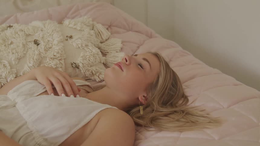 Female lying on her back on a bed sideways with her hand across her chest looks up at the ceiling and then closes her eyes for a moments before opening them again