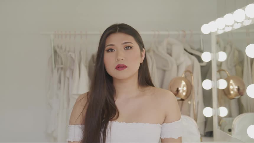 An asian female in a strapless white top stands by a mirror and turns her head to face the camera