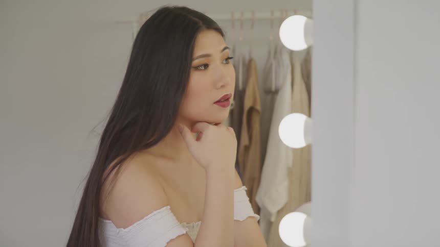 Asian woman with long straight hair and a white dress looks in a mirror in a dressing room